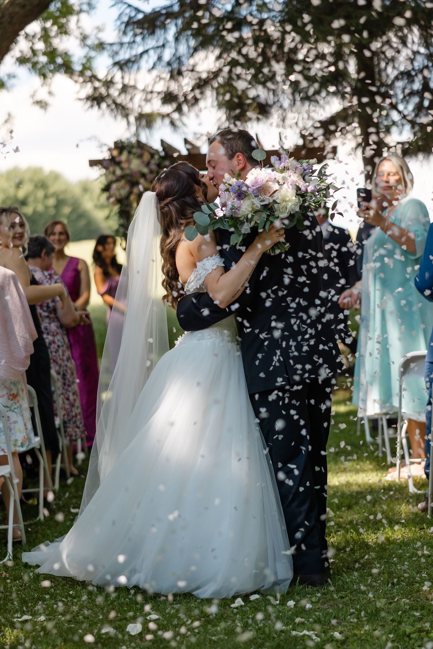 Bride and groom share first kiss at outdoor wedding ceremony as guests toss white flower confetti.