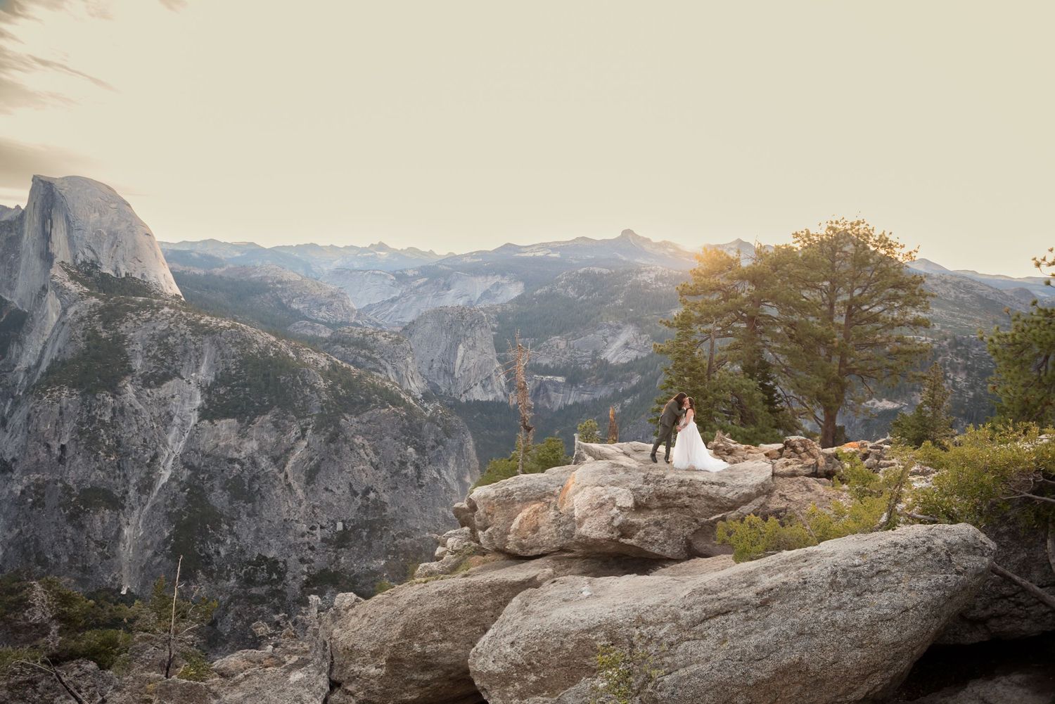 Bride and groom stand on rocky overlook at Yosemite with Half Dome and golden sunset mountain views.