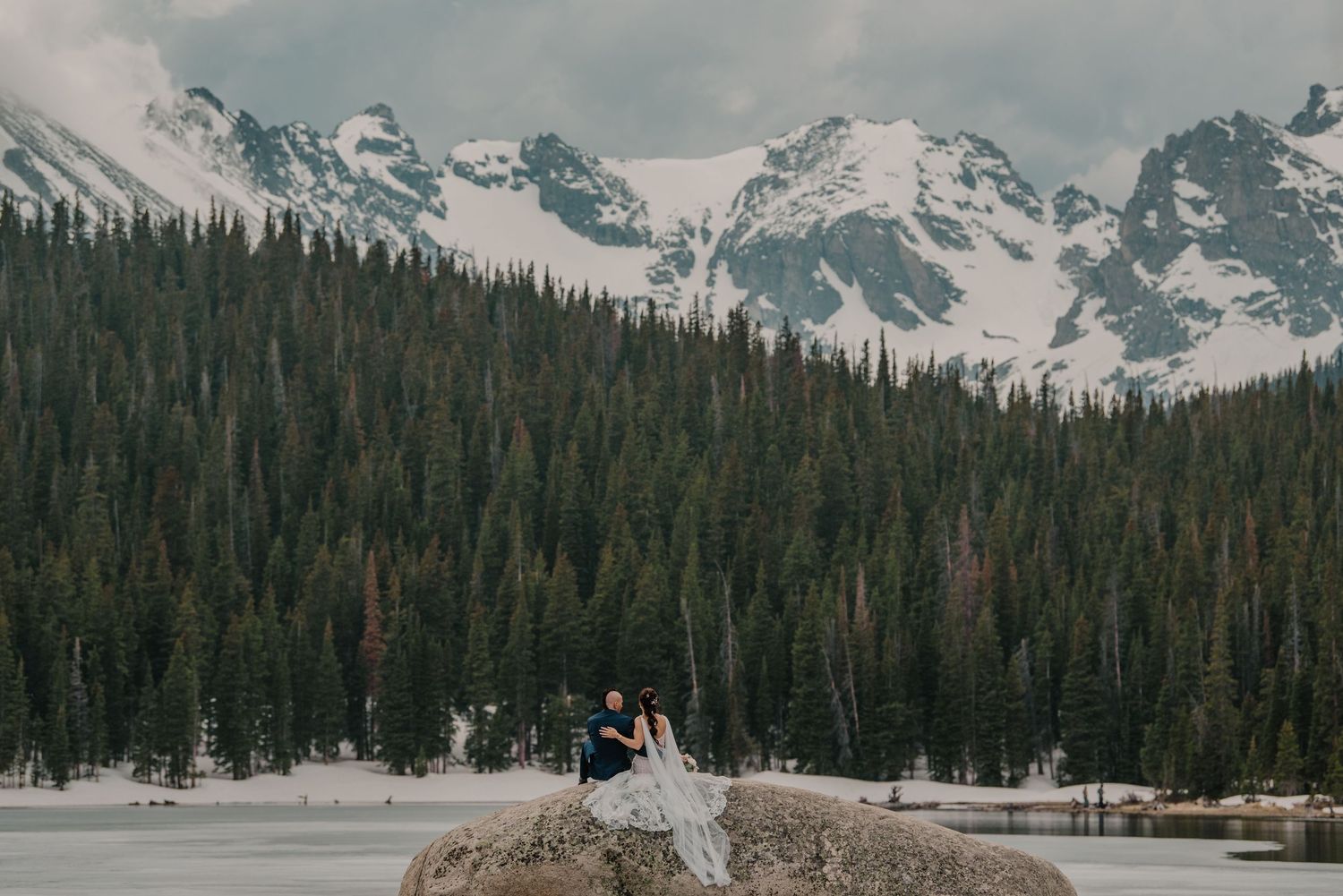 Two people share an intimate moment at a snow-covered mountain lake surrounded by evergreen forest and rugged peaks.