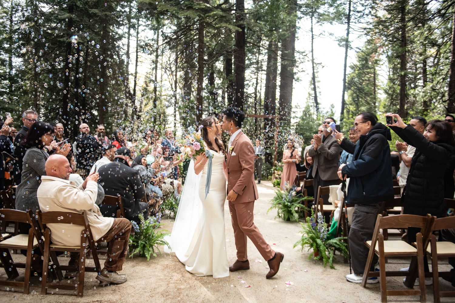 Couple shares first kiss as newlyweds during outdoor forest wedding ceremony with guests tossing confetti.