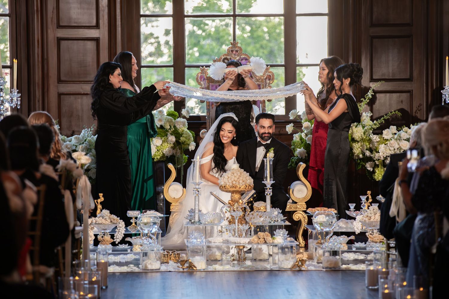 Bride and groom seated at Persian Sofreh Aghd wedding ceremony table with ornate decorations and attendants holding fabric.