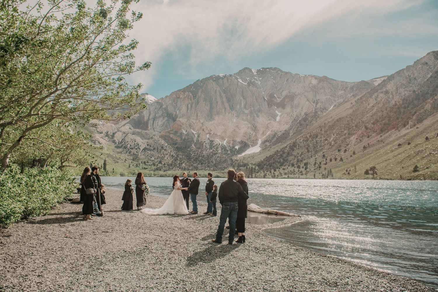 Outdoor wedding ceremony on rocky lakeshore with mountain backdrop, bride and groom surrounded by guests under blue sky.