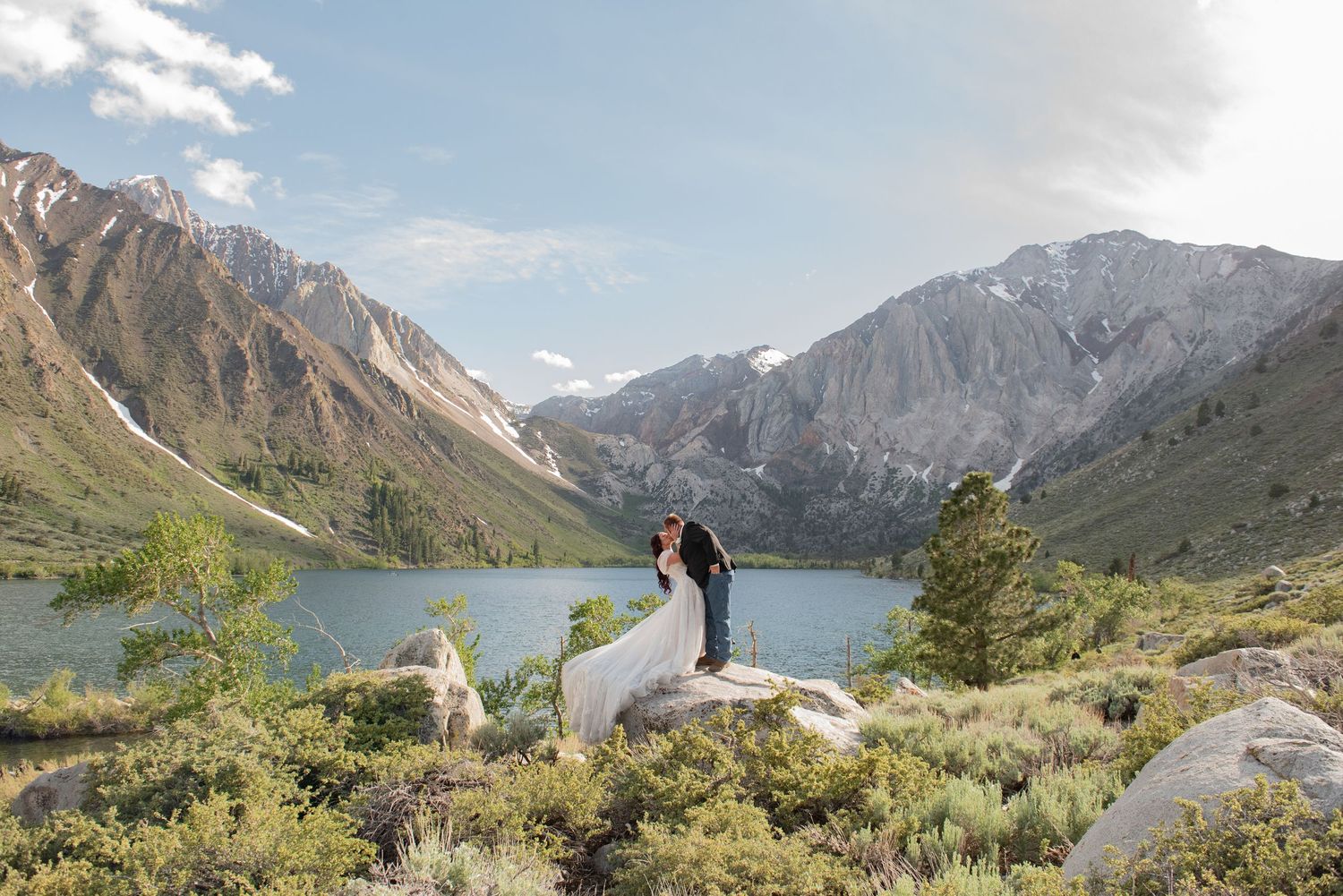 Couple in wedding attire embracing on rocks at Convict Lake with stunning Sierra Nevada mountains backdrop.