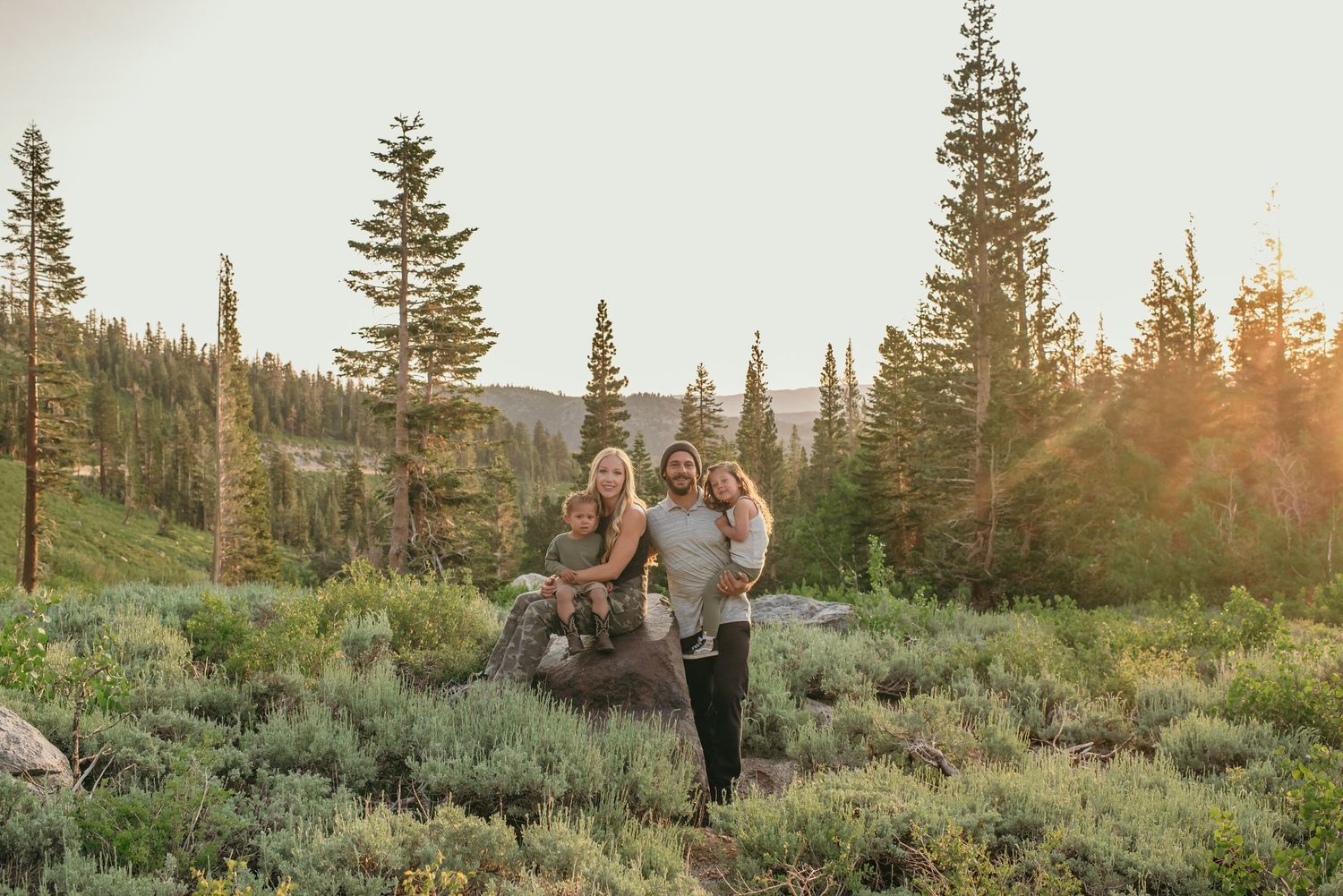 A couple stands together in a mountain meadow at sunset among evergreen trees and sage brush.