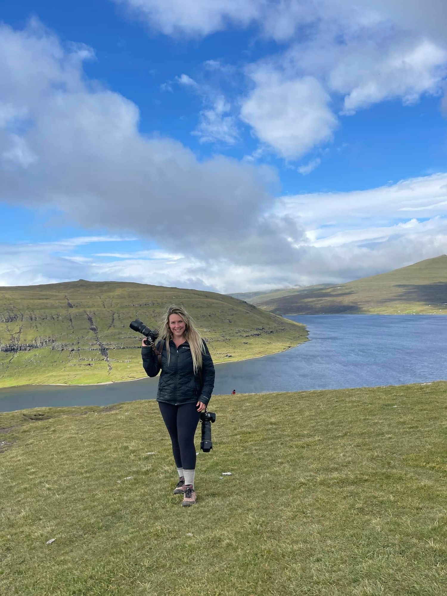 Woman photographer with camera gear stands on green hillside overlooking scenic lake and rolling hills under blue sky.