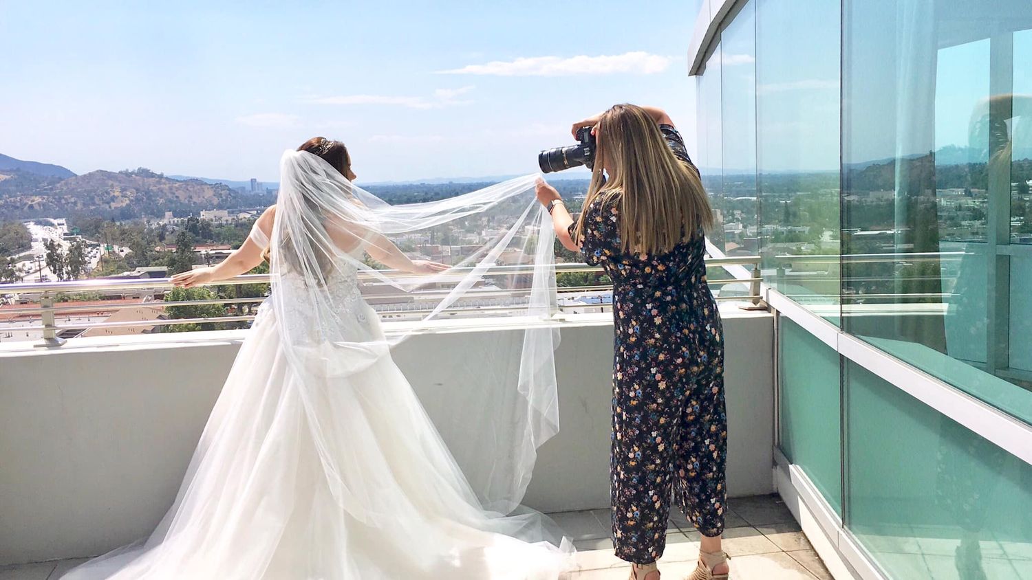 Photographer capturing bride in white wedding gown and veil on rooftop balcony with city and hills view.