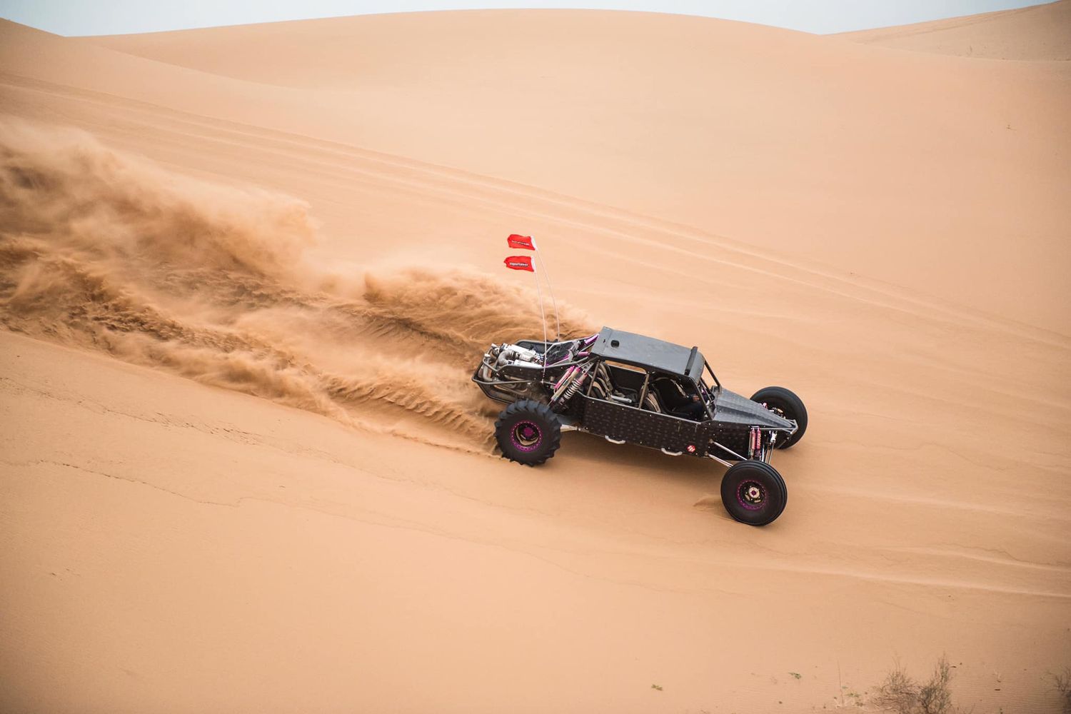 A black off-road buggy races across sandy desert dunes, kicking up dust with a red flag visible in the background.