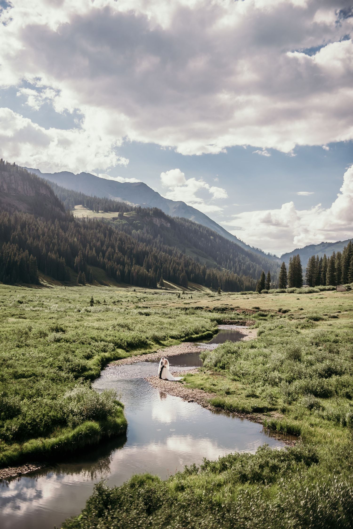 A couple stands by a winding mountain stream in a lush green valley surrounded by pine forests and dramatic cloudy skies.