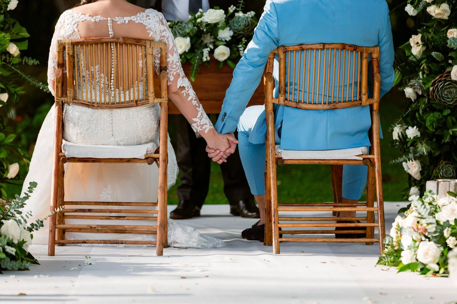 Pareja de novios tomados de la mano durante la ceremonia vista desde atrás.
