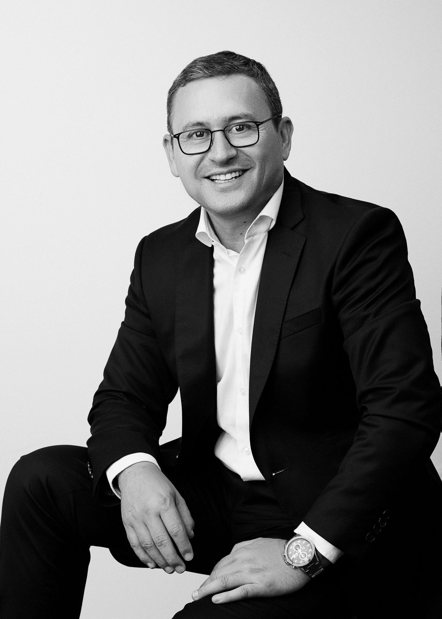 Professional business portrait in black and white showing person in suit and glasses sitting casually against light background.