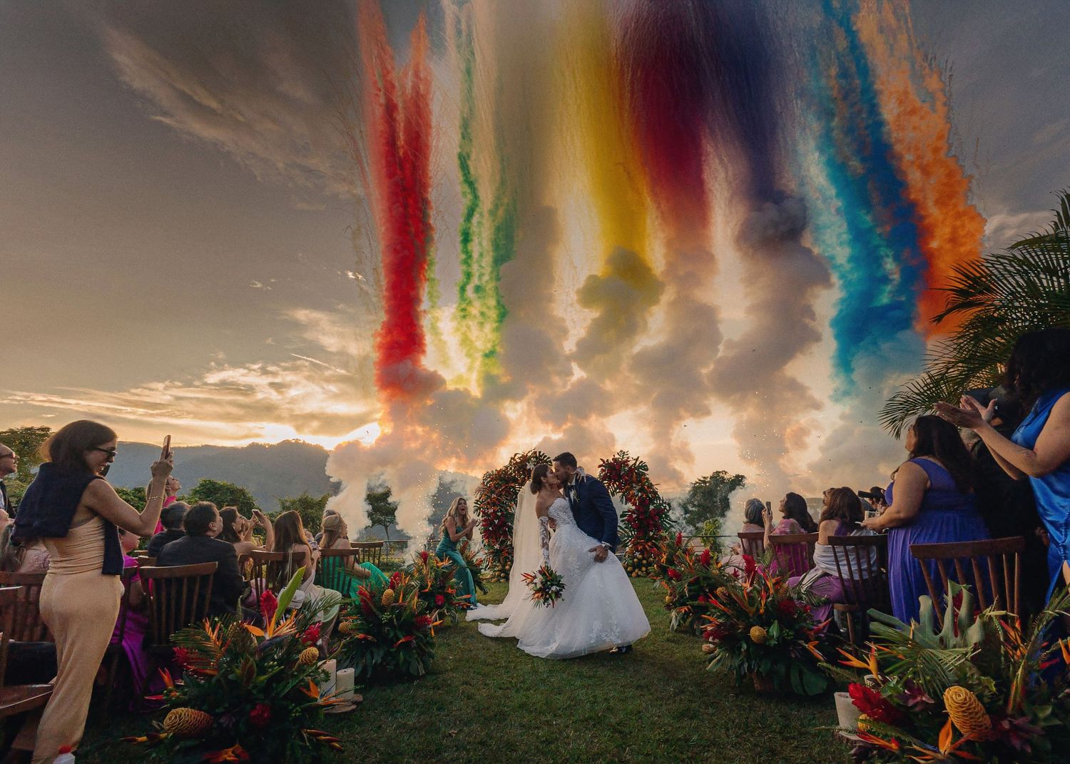 Colorful smoke bombs create vibrant trails against a sunset sky during an outdoor celebration.