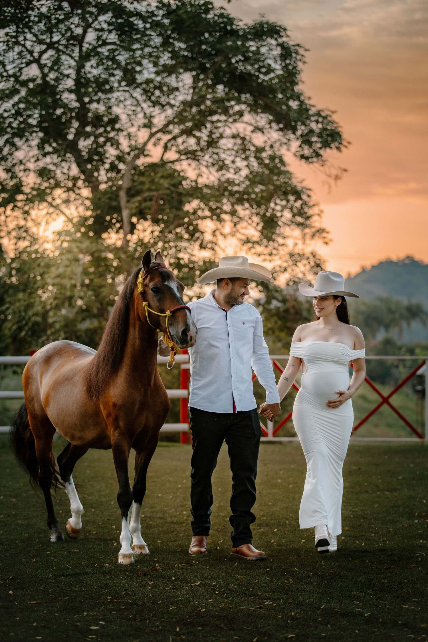 A couple in western attire stands with a brown horse in a sunset pasture during a countryside photoshoot.