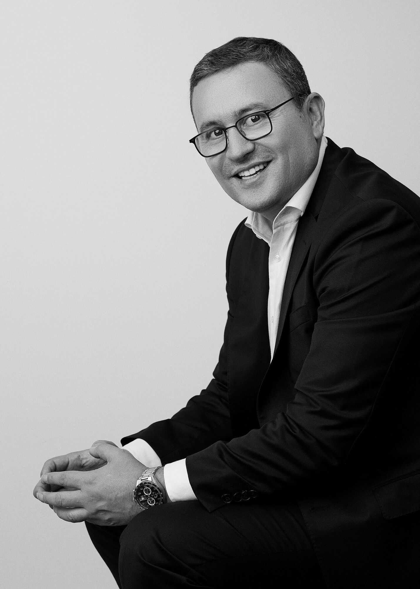 Retrato en blanco y negro de hombre profesional sonriendo con gafas sentado con manos juntas en Pereira — John Palacio