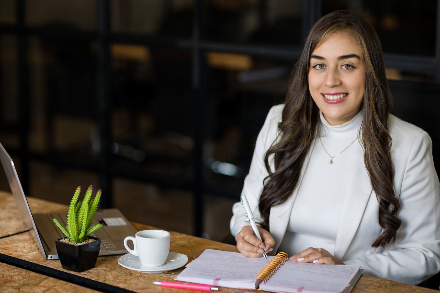 Mujer profesional sonriendo escribiendo en una agenda en un escritorio con portátil y café en una oficina de Pereira — John Palacio