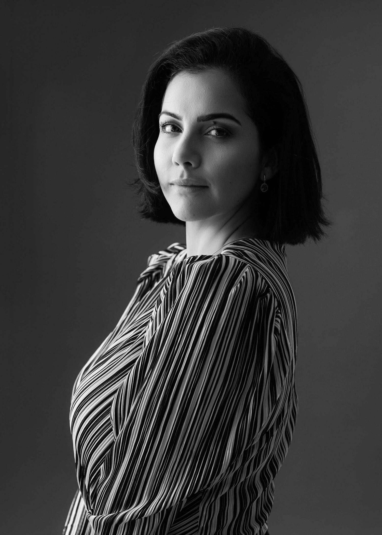 Black and white portrait of someone with shoulder-length dark hair wearing a striped blouse in dramatic lighting.