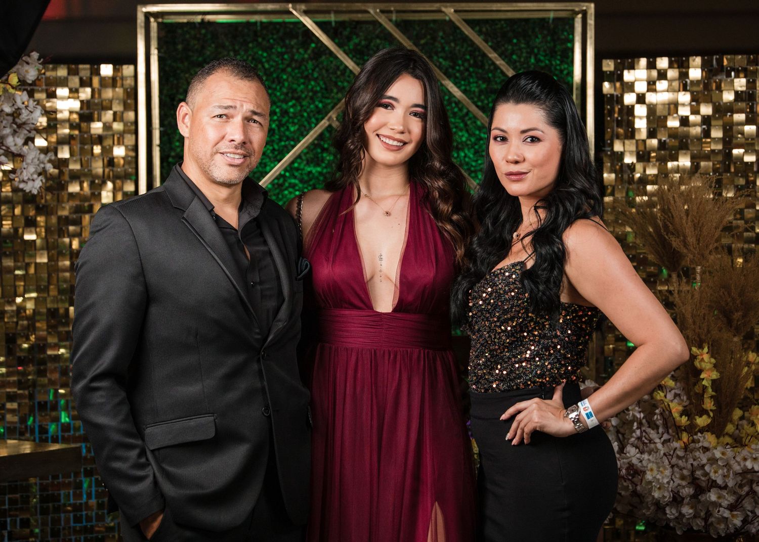 Three people pose together at an elegant event against a backdrop of gold sequins and greenery.