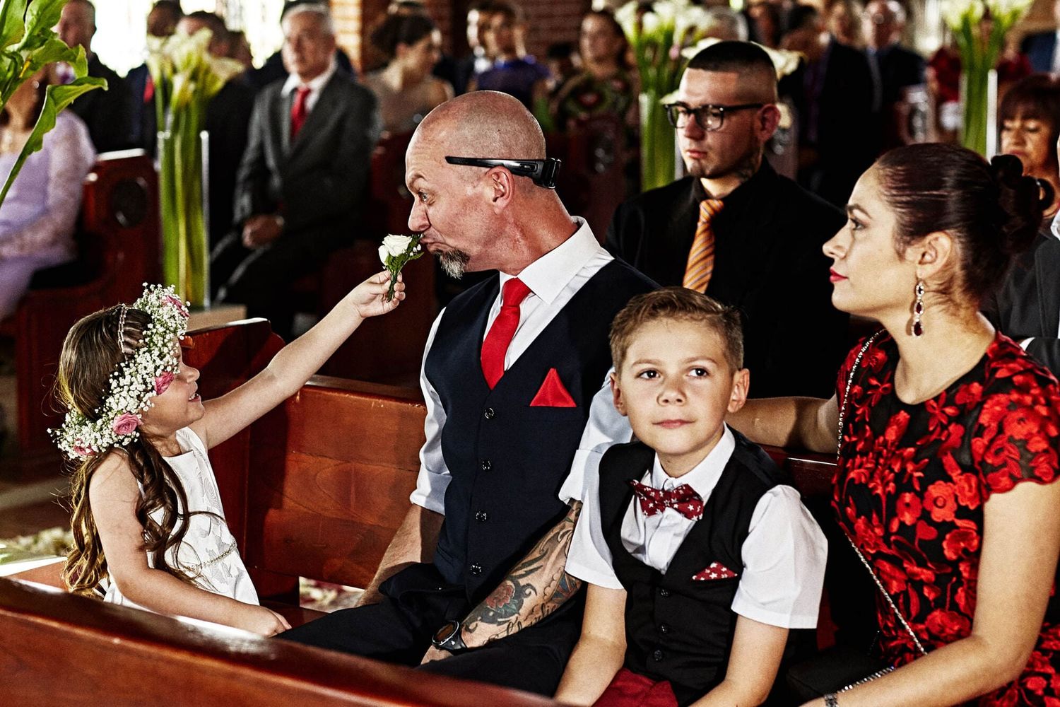 Niña con corona de flores jugando con hombre durante ceremonia de boda, rodeados de familia.