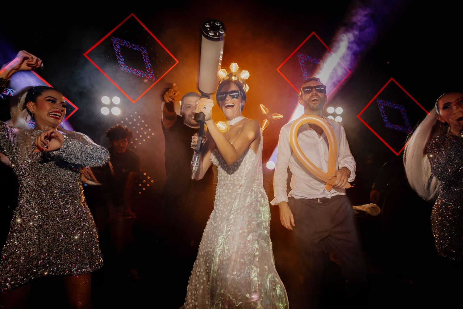 Novia con vestido iluminado festejando en pista de baile con invitados y luces de fiesta.