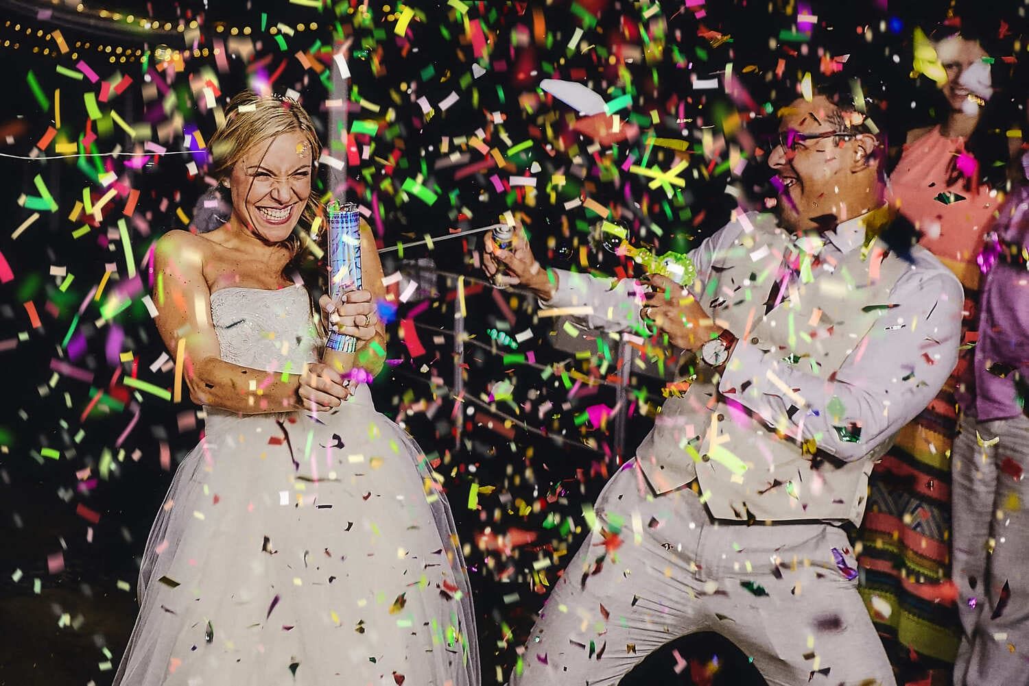 Novios riendo entre confeti durante la fiesta de bodas, ambiente alegre y colorido.