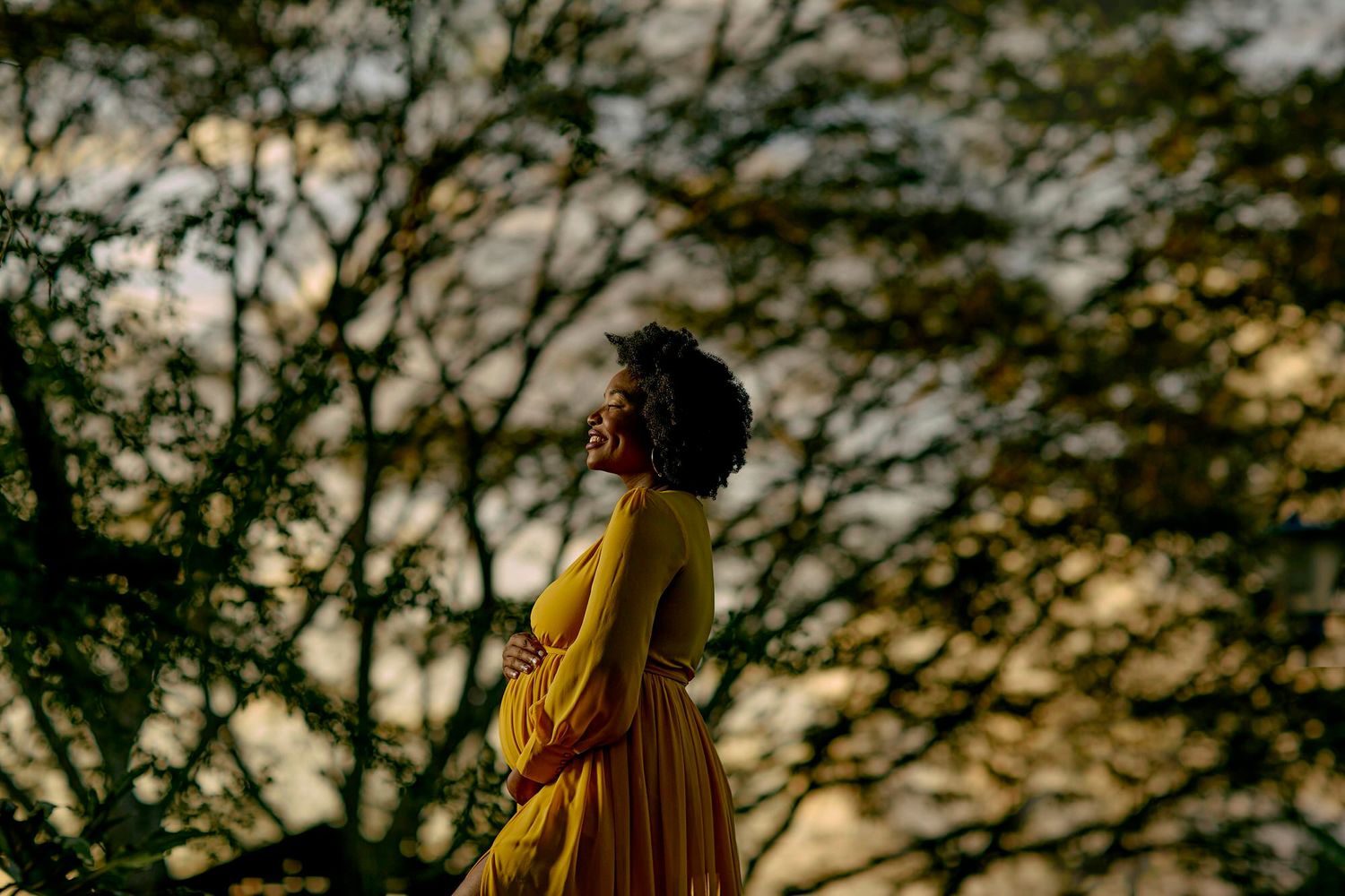 Figure in golden yellow dress stands in silhouette against a sunset-lit forest backdrop with bokeh trees.