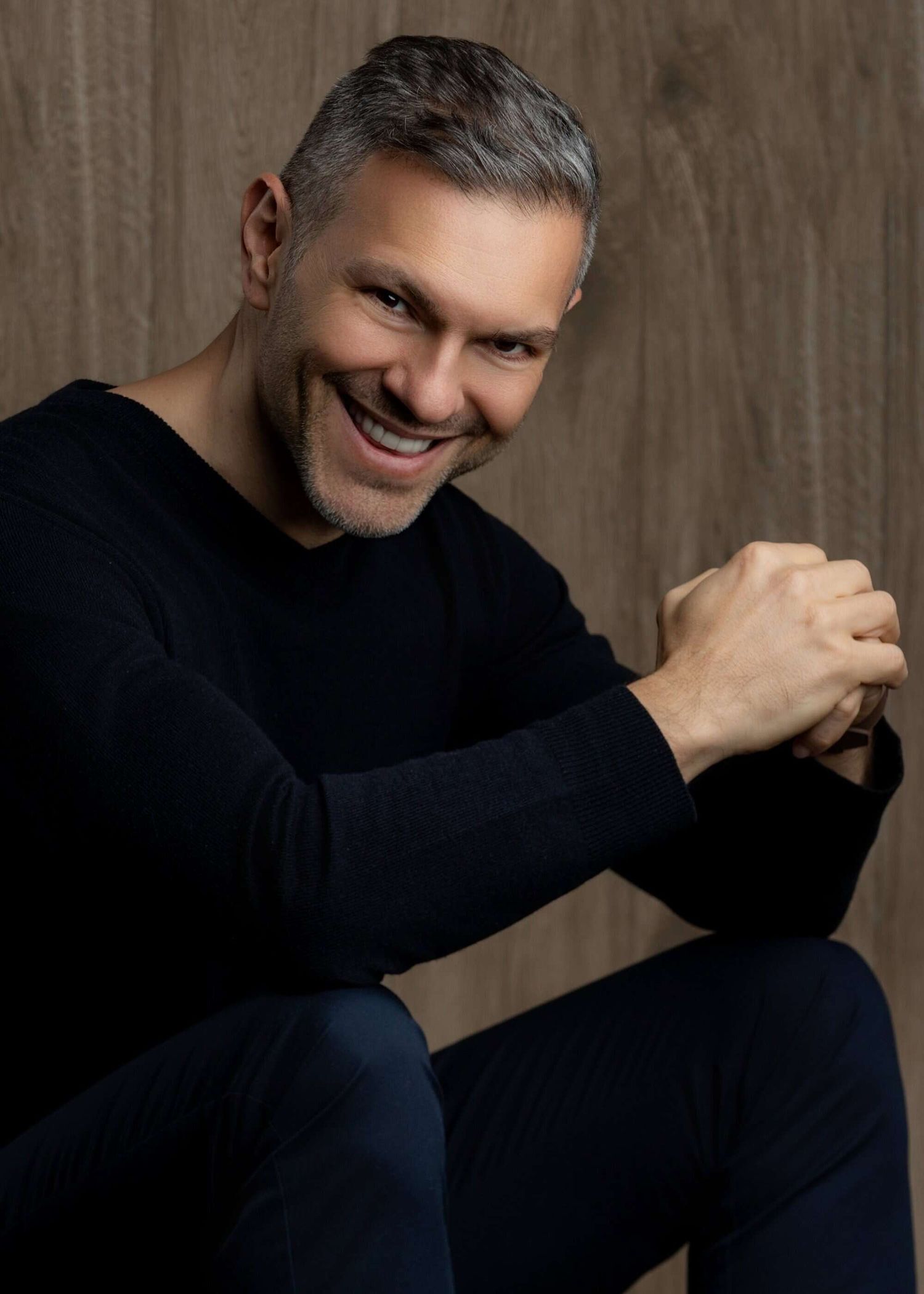Professional portrait of someone in a black sweater sitting with clasped hands and smiling warmly against a brown backdrop.