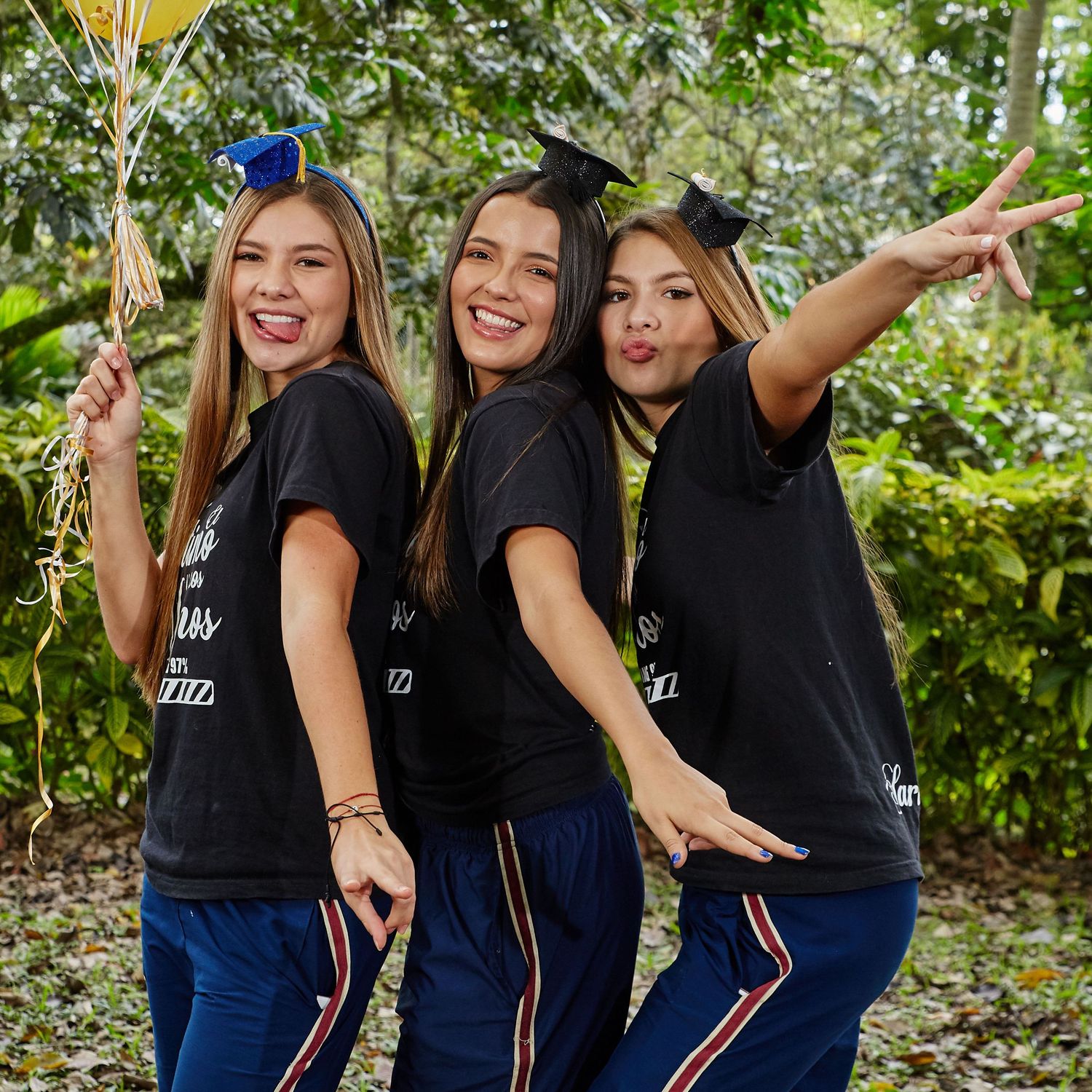 Three friends in black tops and jeans pose playfully together outdoors in a garden setting.