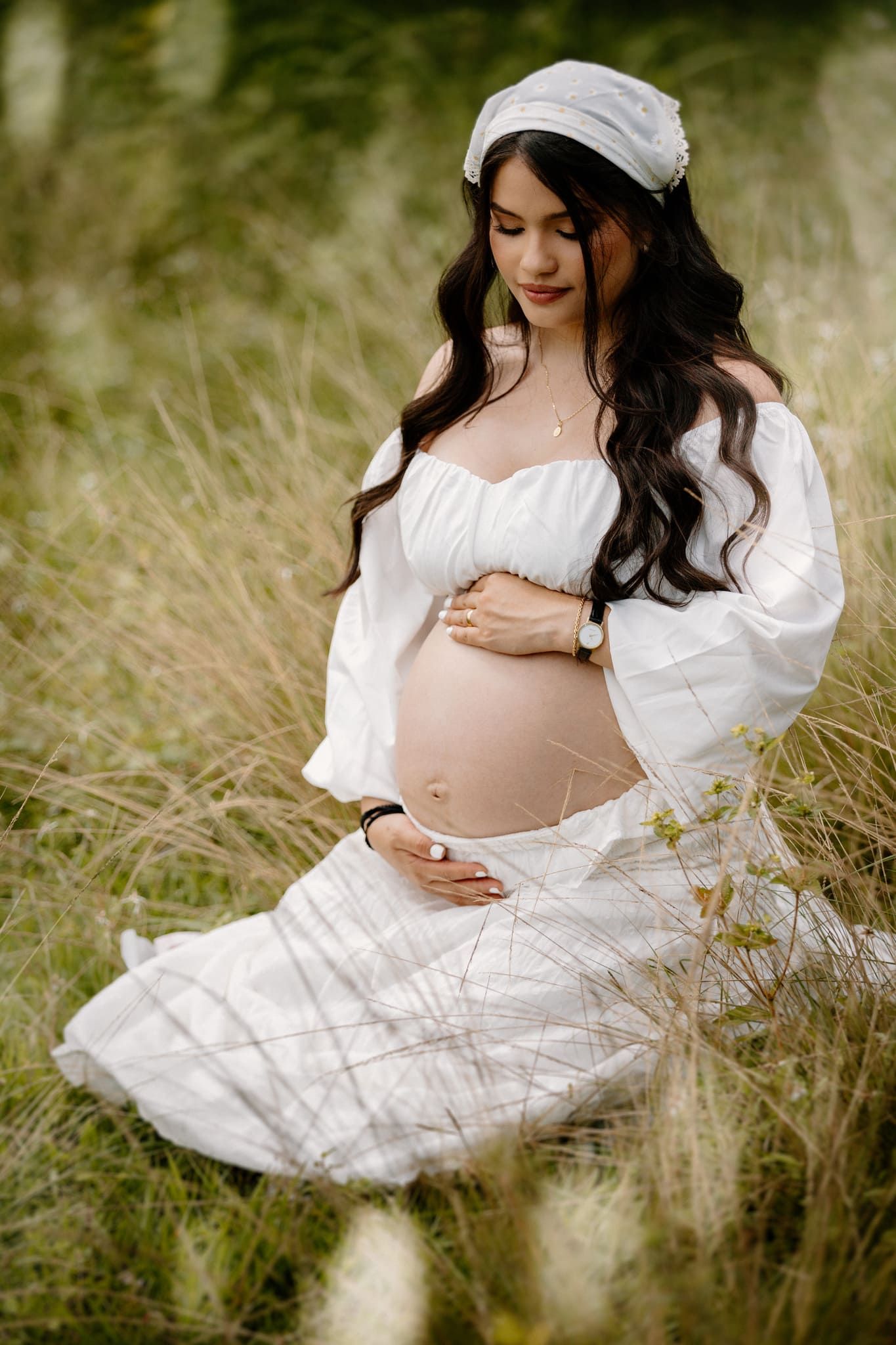 mujer embarazada vestida de blanco sentada en el pasto mirando su barriga en sesion de fotos de maternidad por john palacio