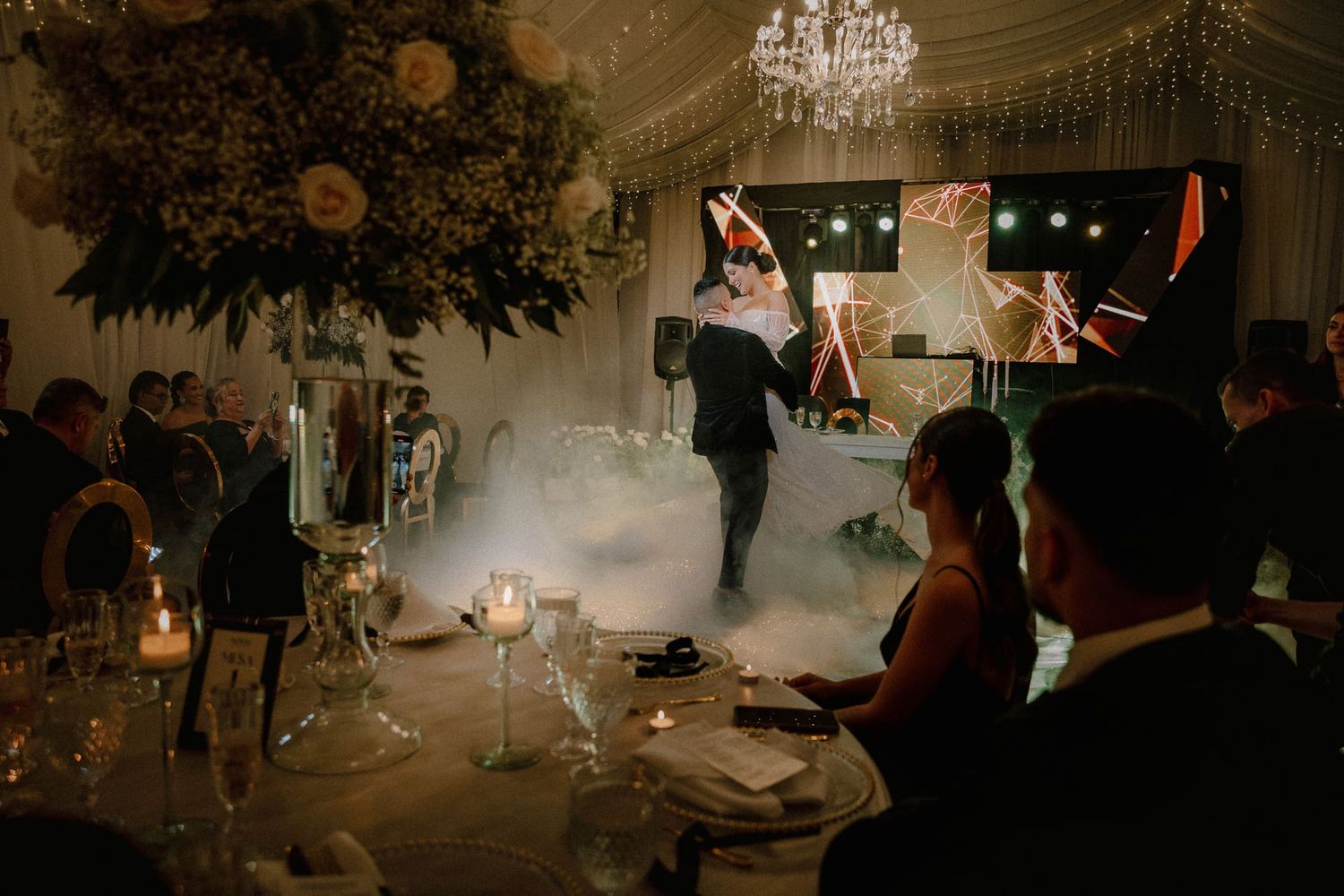 Novios bailan su primer vals rodeados de invitados y luces en una elegante recepción de matrimonio.