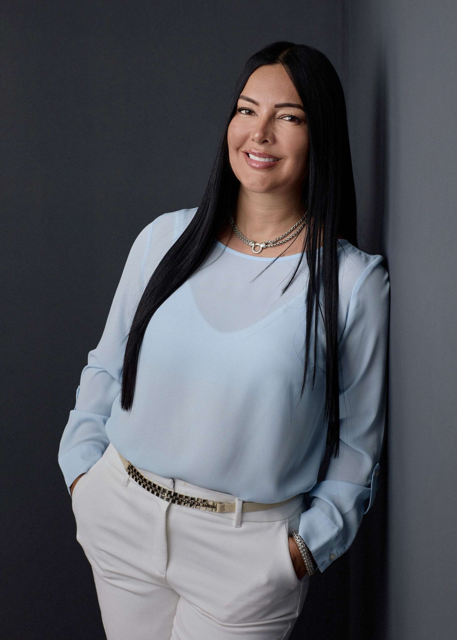 Mujer profesional de cabello negro largo en blusa azul recostada en una pared sonriendo en Pereira — John Palacio