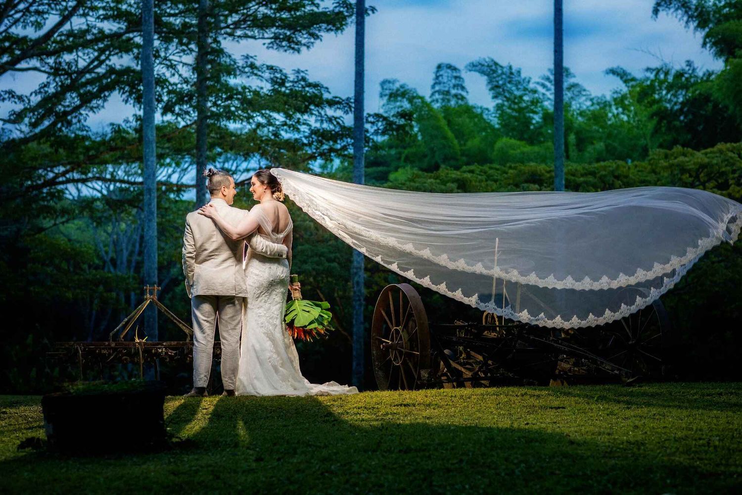 A couple shares a romantic moment outdoors as a wedding veil flows dramatically in the breeze during their tropical garden ceremony.
