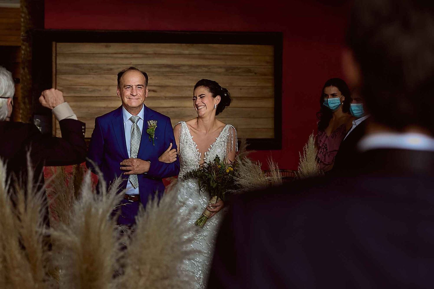 Novia sonriente entrando a la ceremonia del brazo de un familiar, rodeada de invitados.