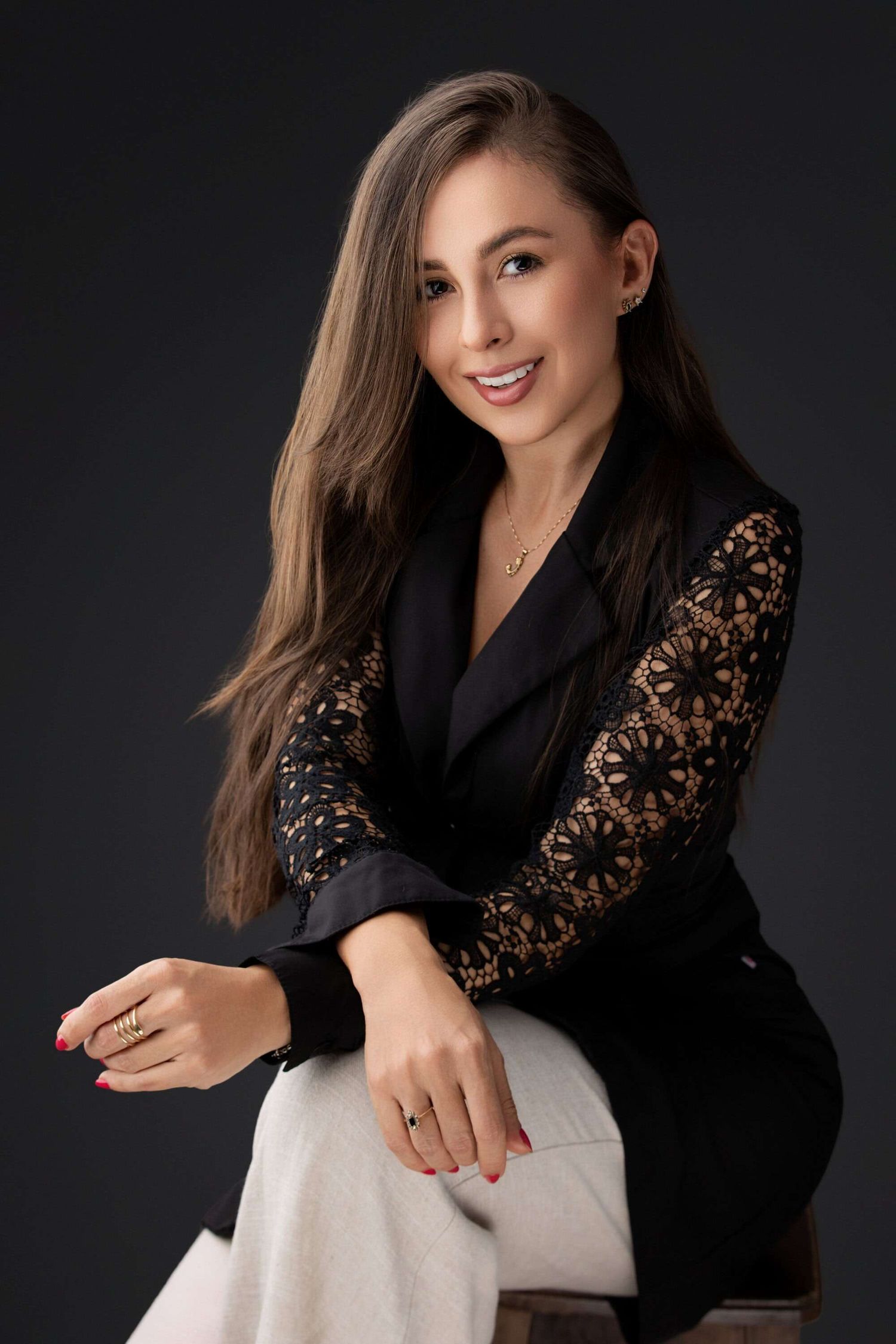 Professional portrait of someone in a black blazer with lace sleeves seated on a light colored chair against dark background.