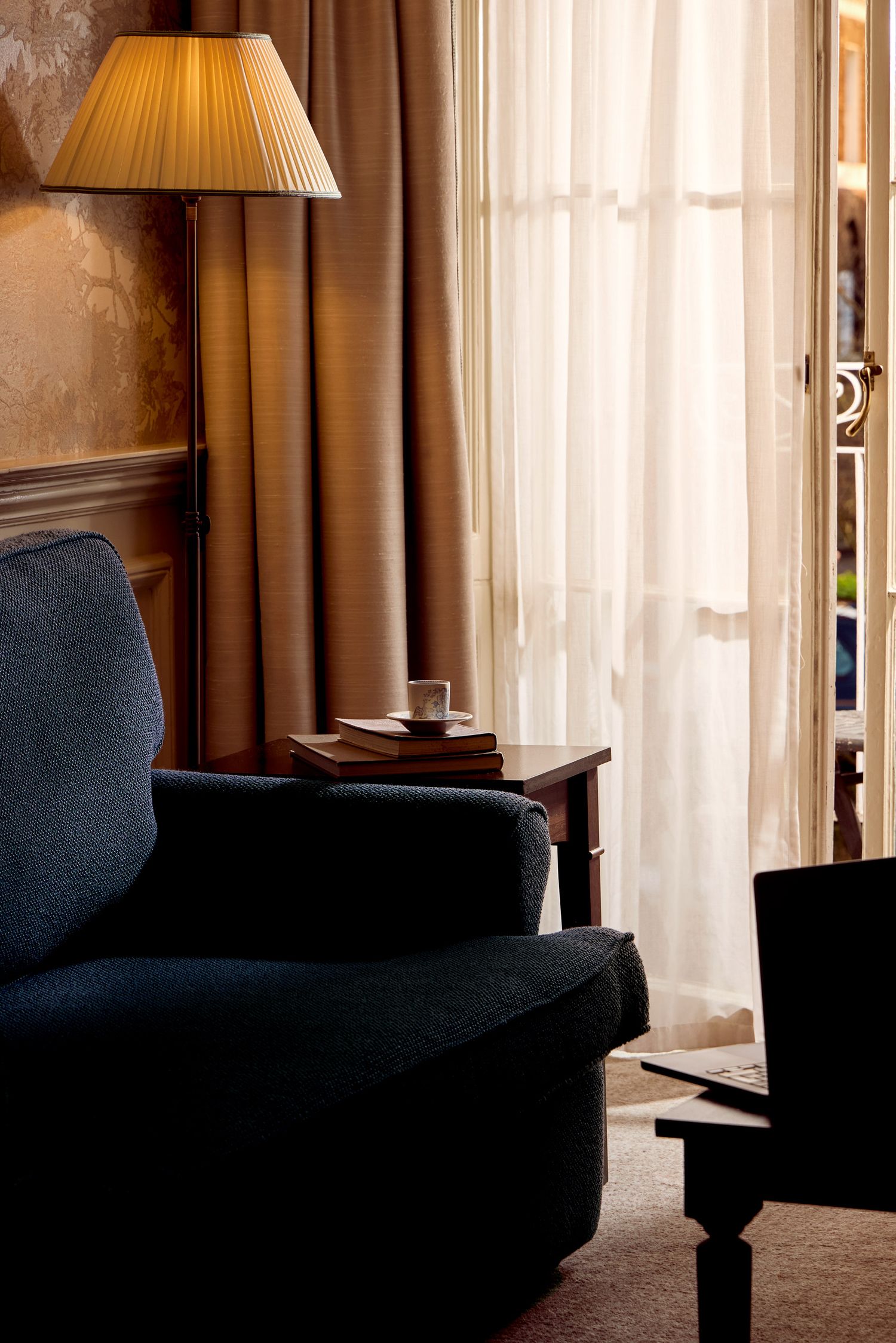 Elegant hotel room in London with table lamp, sheer curtains, and dark armchair in warm ambient lighting.