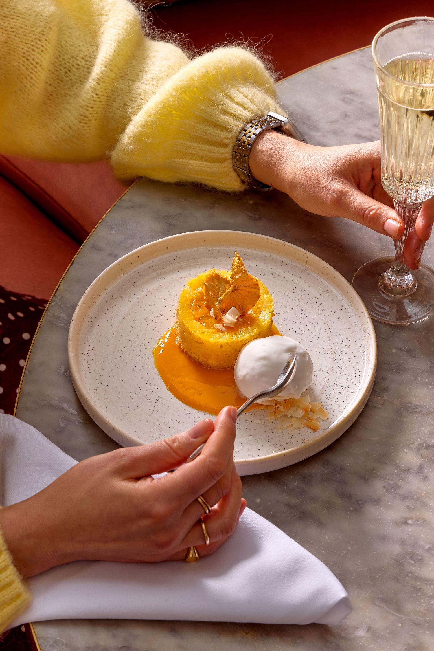 Hands holding utensils over a dessert plate with an orange poached pear and cream, alongside a champagne glass in a London restaurant