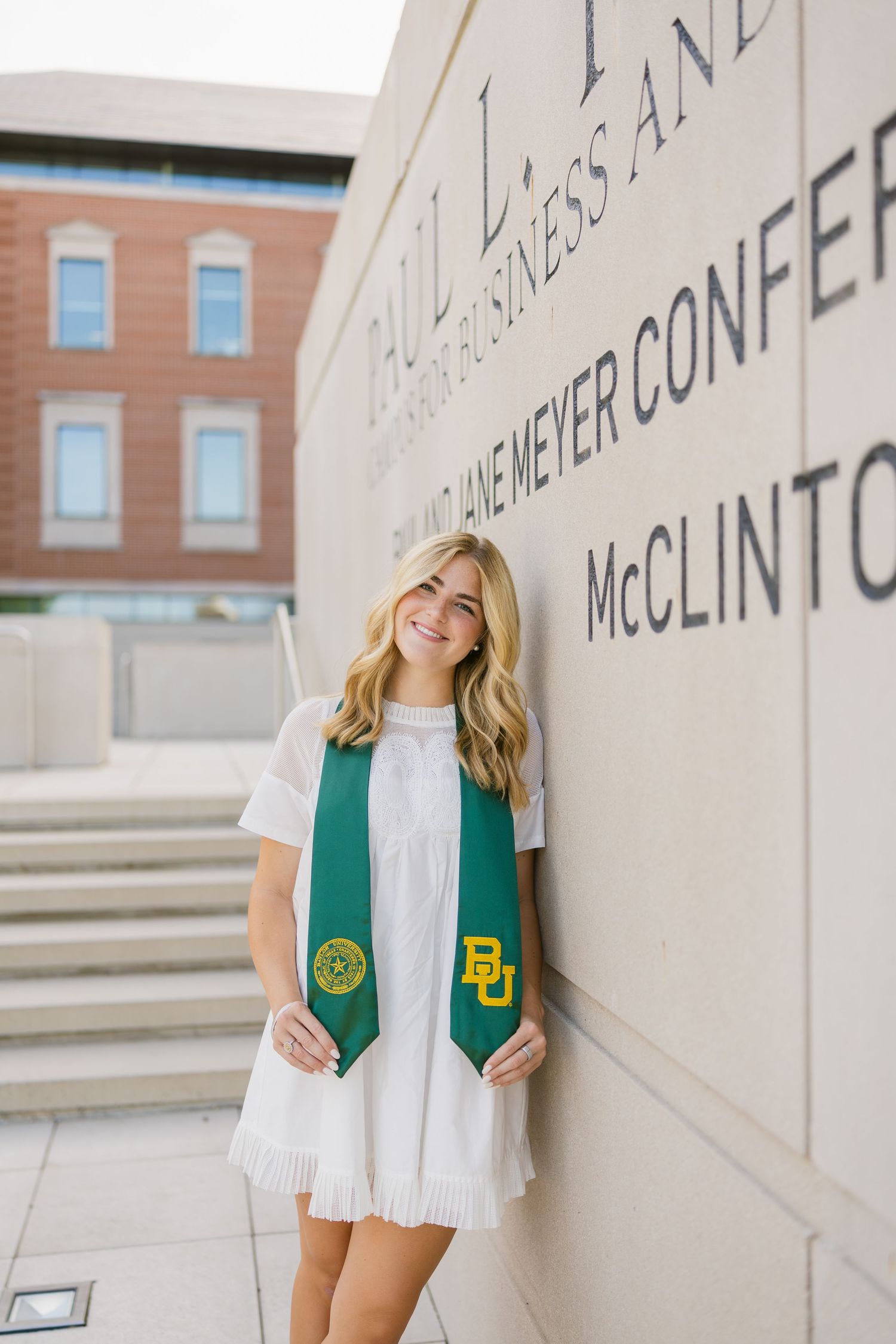 Blonde graduate in white dress and green BU stole leans against a Baylor University Hankamer School of Business wall.