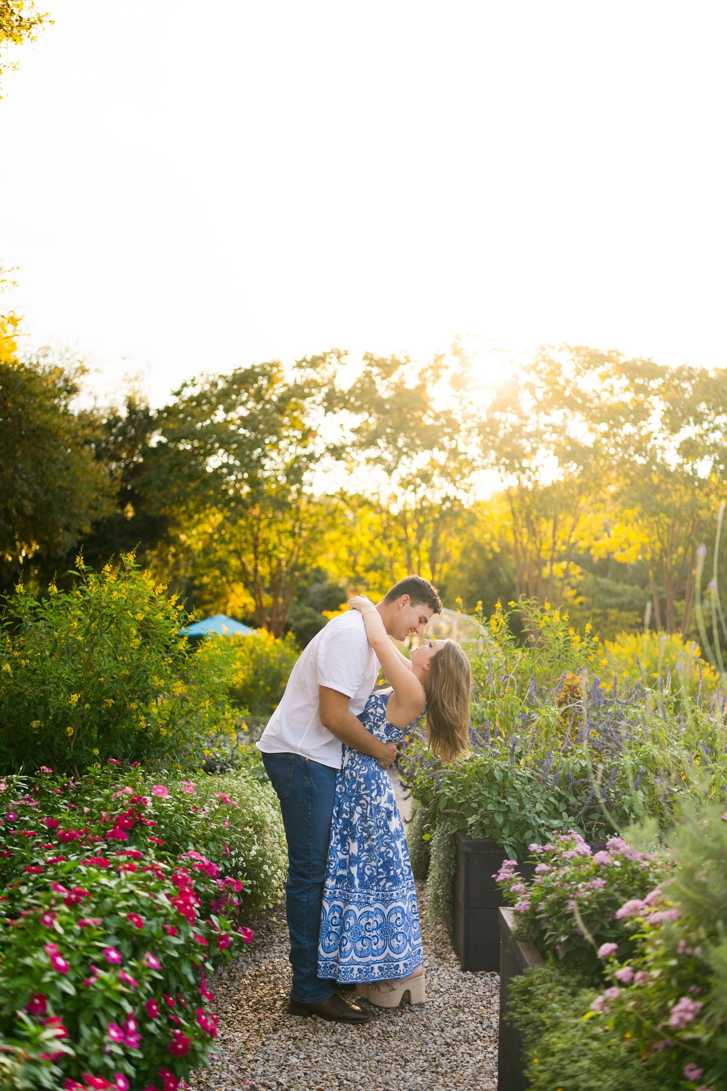 A couple shares a romantic kiss in a vibrant flower garden bathed in golden sunset light.