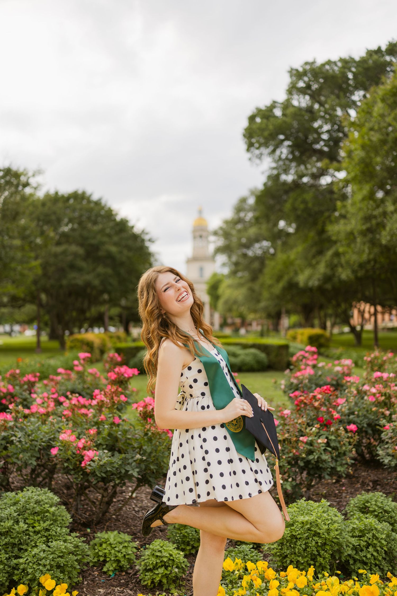 Happy young woman in polka dot dress poses playfully in a flower garden at Baylor University.