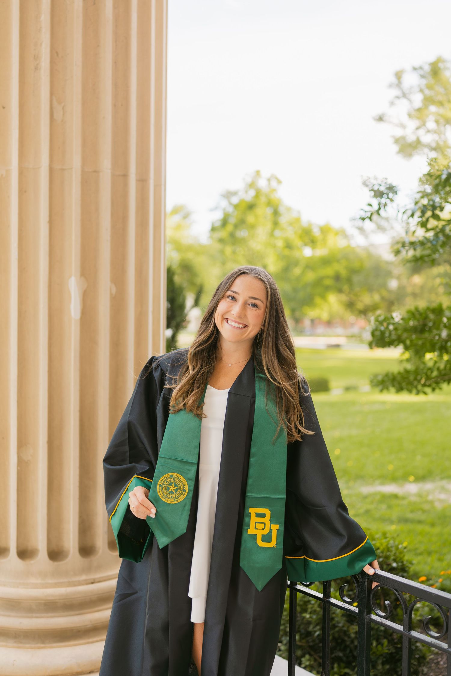 Smiling Baylor University graduate in black cap and gown with green BU stole poses by a stone column.