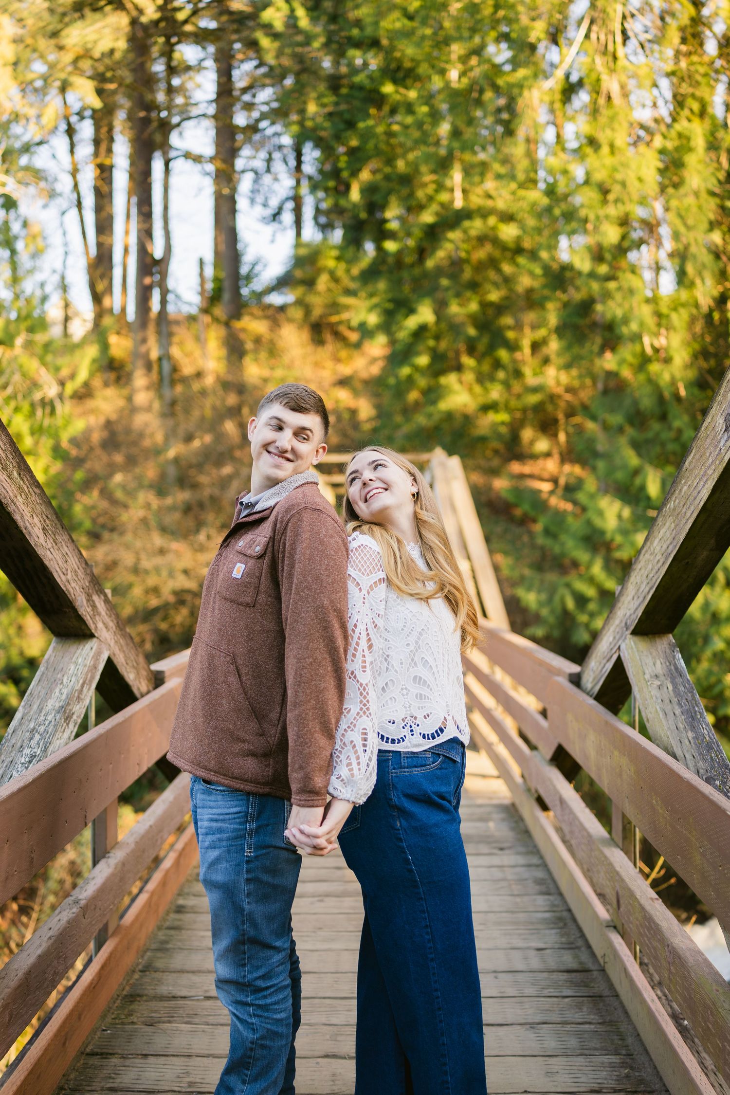 A happy couple holds hands and smiles on a wooden bridge surrounded by colorful autumn foliage.