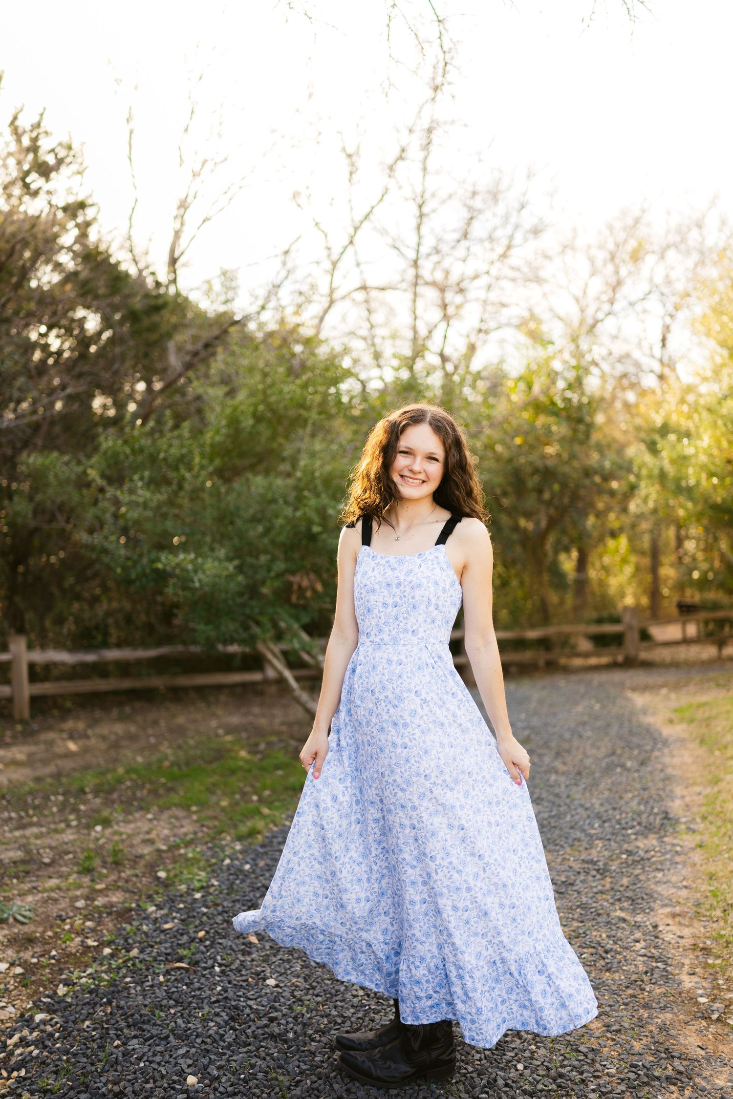 A young woman in a blue-and-white floral maxi dress smiles as she stands on a gravel path outdoors, twirling it.