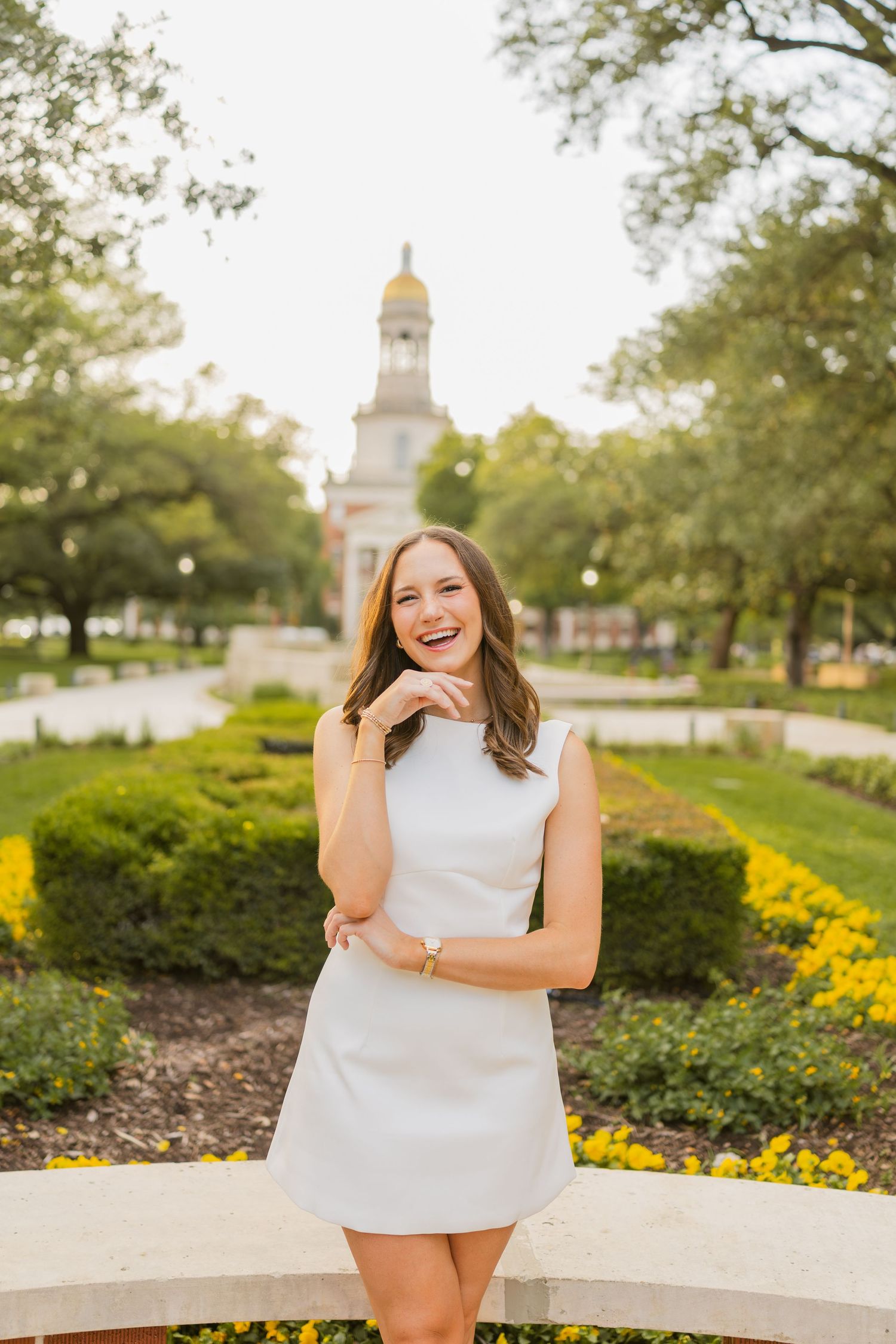 Laughing young woman in a white mini dress poses in a garden with Baylor University's Pat Neff Hall in the background.