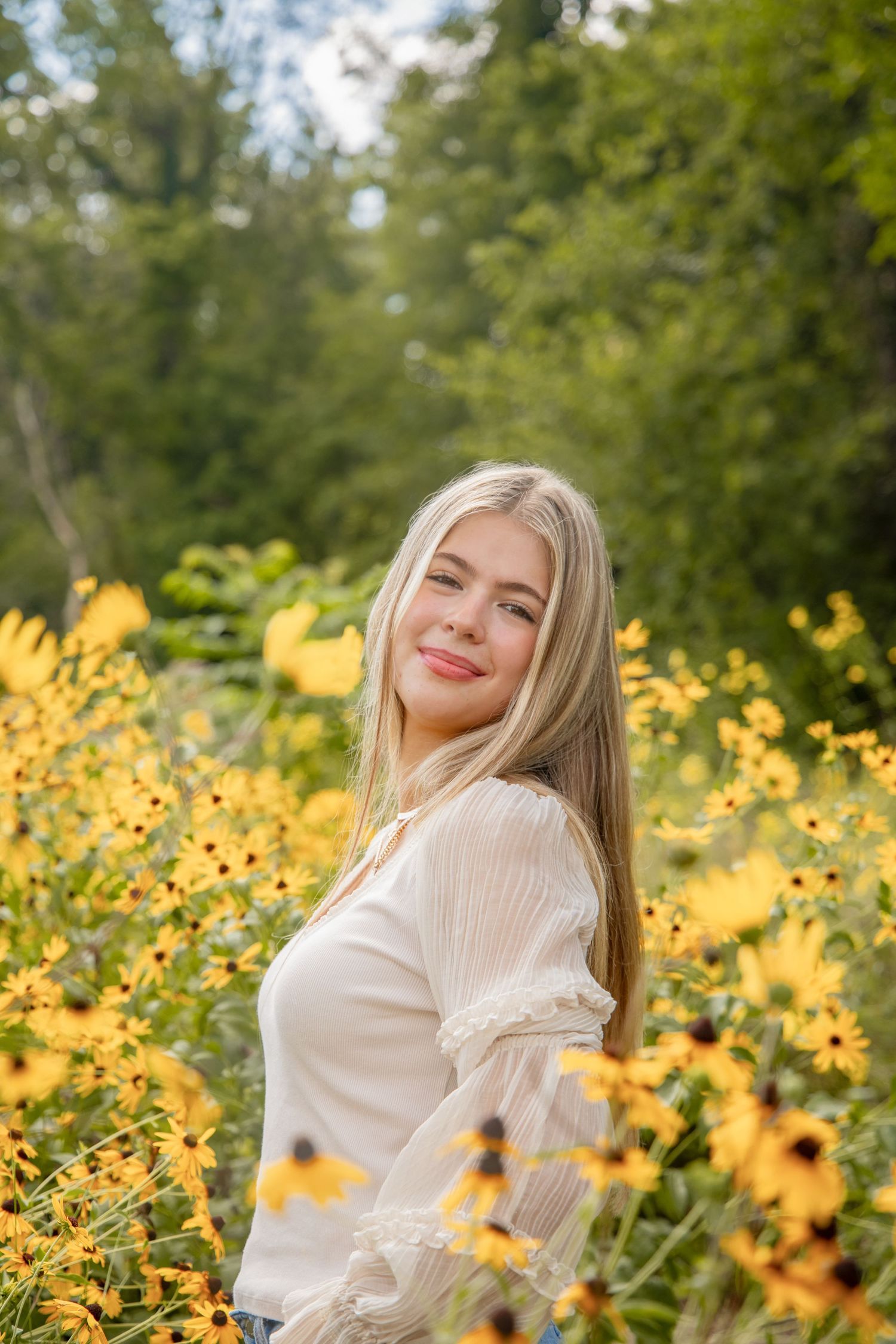 Blonde teenage girl in white blouse smiles softly while surrounded by yellow wildflowers in a lush garden.
