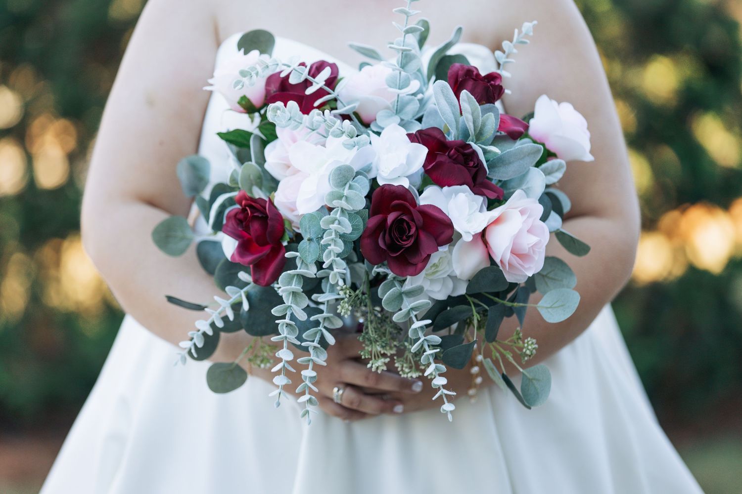 A burgundy and white rose wedding bouquet with eucalyptus leaves held against a white wedding dress in natural outdoor lighting.