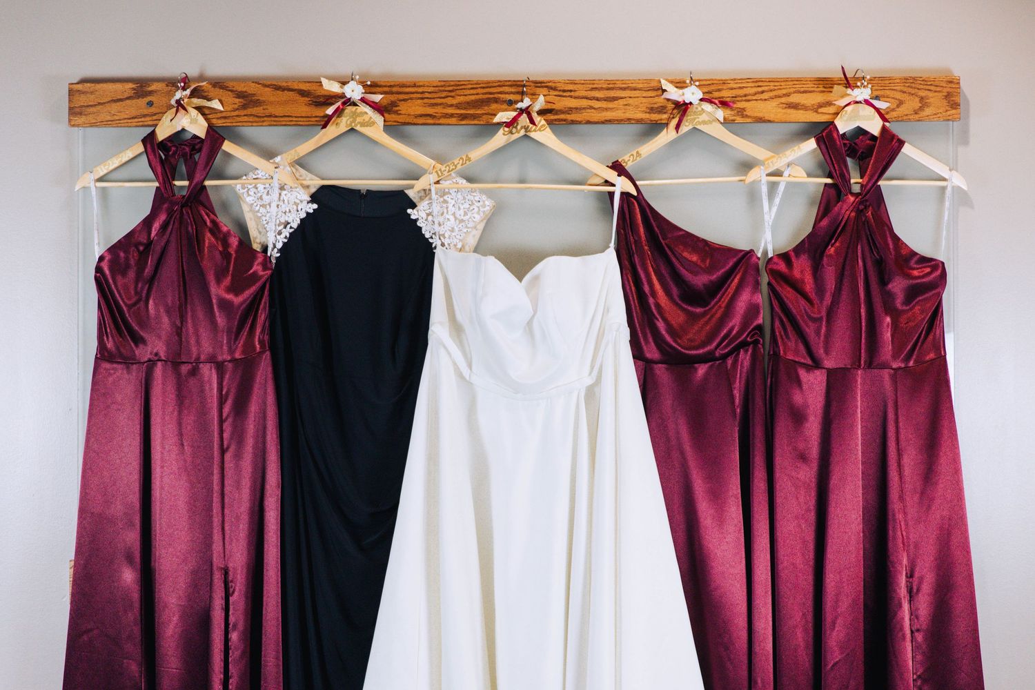 Wedding gown and burgundy bridesmaid dresses hanging on wooden rack against gray wall.