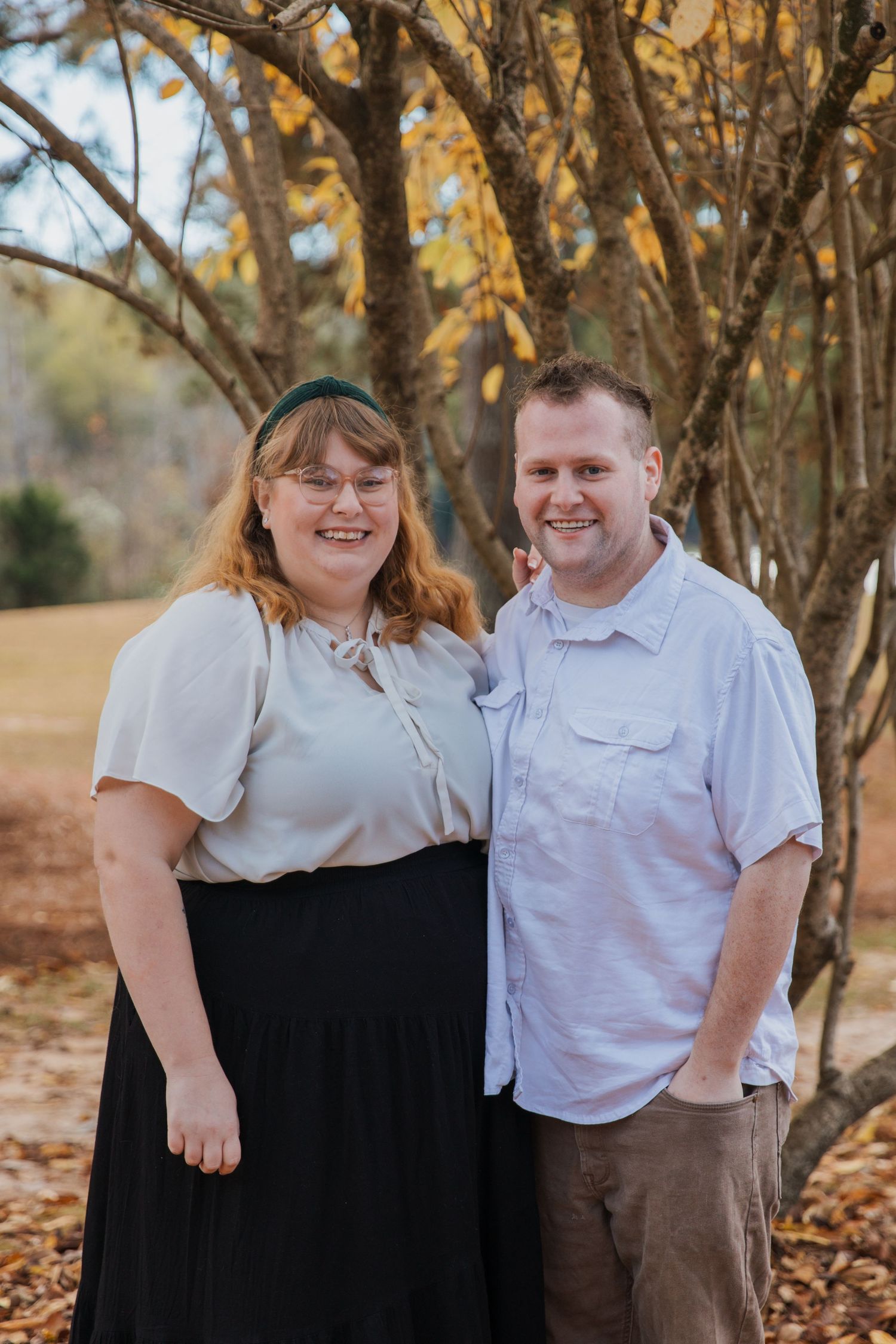 A couple poses together outdoors in coordinating white tops against an autumn backdrop with golden leaves.