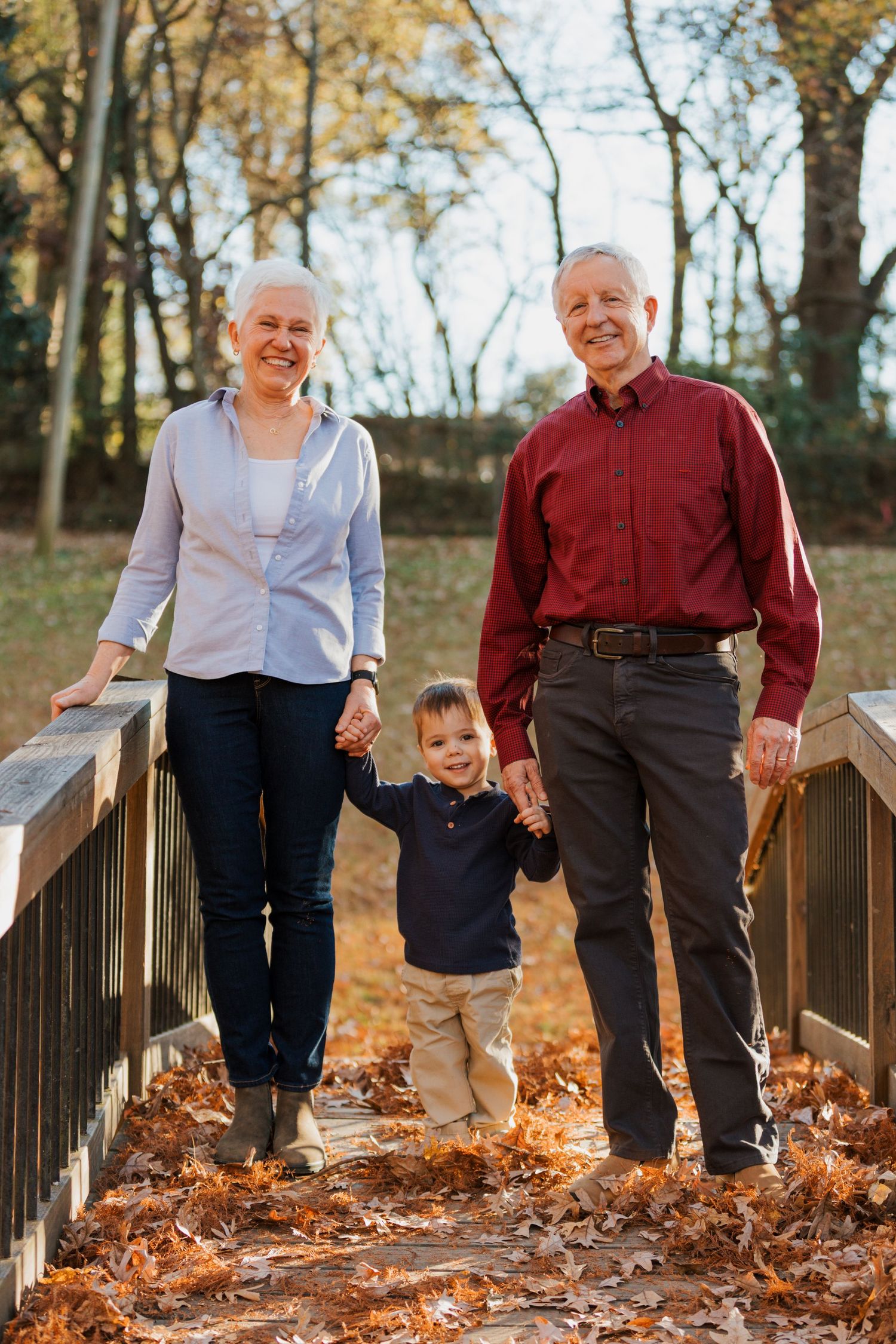 Three generations walking together on a wooden bridge covered in autumn leaves on a fall day.