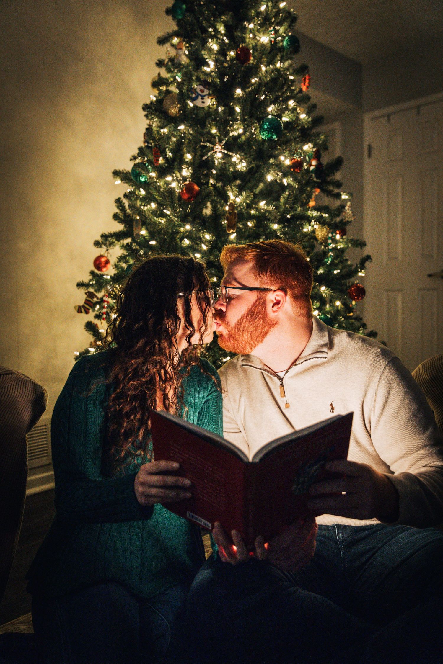 A couple shares an intimate moment while reading together by a decorated Christmas tree in a dimly lit room.