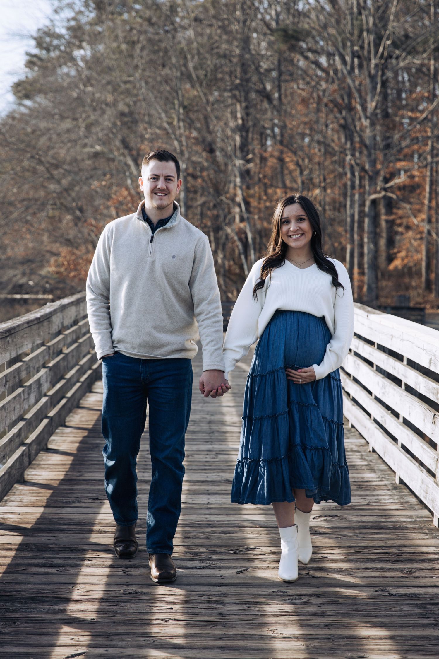 Couple walking hand in hand on wooden bridge during autumn, with woman wearing blue tiered skirt and white boots.