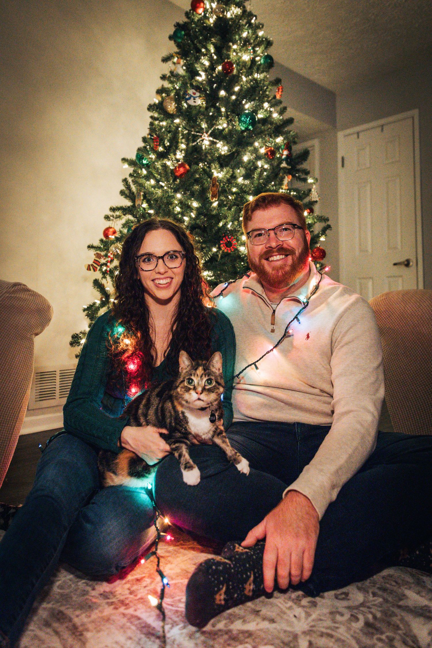 Couple sitting on couch with Christmas tree and string lights creating festive holiday atmosphere.