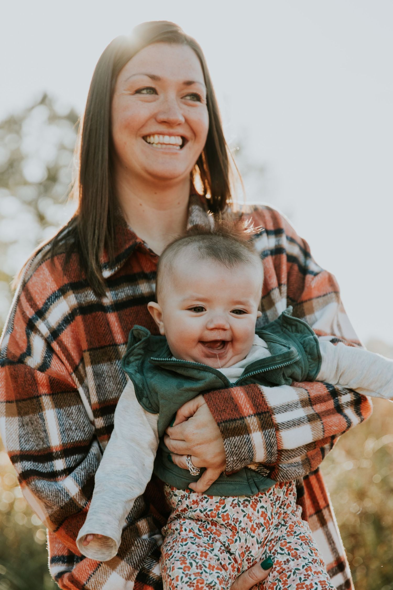 A parent in a plaid flannel shirt holds a smiling baby wearing a green jacket and floral pants outdoors.